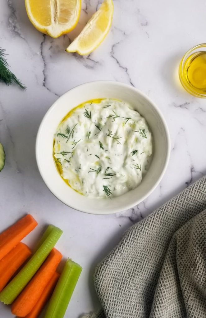 bowl of homemade tzatziki sauce with fresh dill and olive oil on top, small ramekin of olive oil on the top right, lemon wedges on the top left, carrot and celery sticks in the bottom left hand corner, some fresh dill poking out the left side of the frame