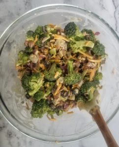 bowl of chopped cooked bacon, diced red onions, grated cheddar cheese, dried cranberries, sunflower seeds and broccoli florets mixed together with a creamy sauce. rubber spatula in bowl