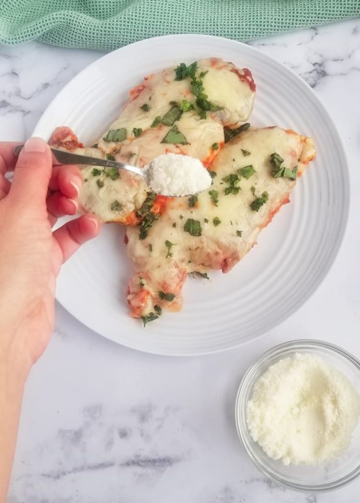 a plate with two cooked chicken breasts topped with melted mozzarella cheese and chopped fresh parsley. A bowl of grated parmesan cheese on the bottom right with a hand holding a spoon of parmesan cheese oven the chicken