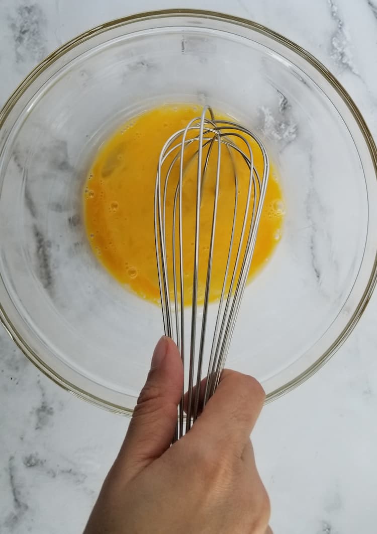 a hand holding a whisk in a glass bowl beating an egg