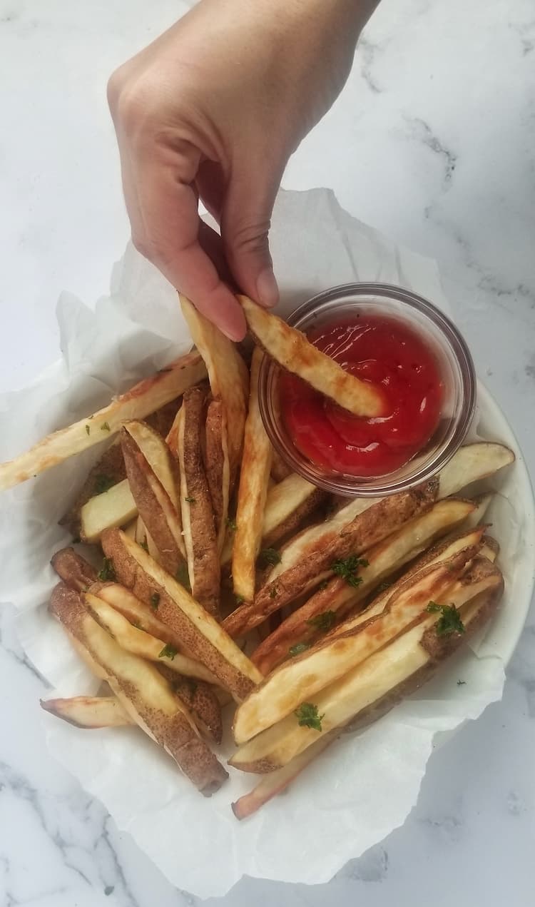bow of air fryer fries with ketchup and chopped parsley, hand dipping a fry into the ketchup