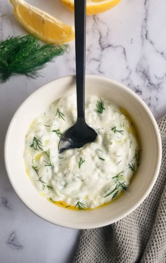 bowl of homemade tzatziki sauce with a black spoon, olive oil on top, fresh dill on top and in the background next to some lemon wedges
