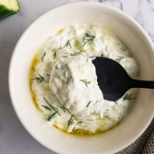 black spoon lifting up some tzatziki sauce over a bowl of the rest, fresh dill and olive oil on top, cucumber barely visible on the top left side