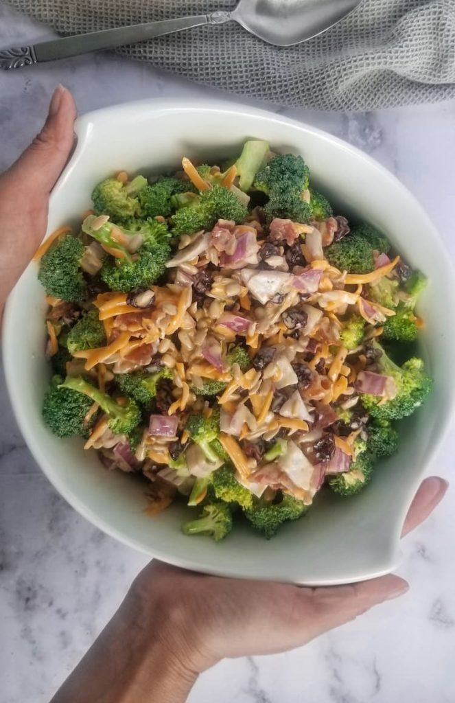 two hands holding a big bowl of broccoli with shredded cheddar cheese, red onions, dried cranberries, sunflower seeds, spoon and grey cloth behind bowl