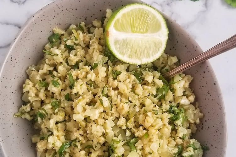 bowl of riced cauliflower mixed with chopped cilantro, two spoons in the bowl, half lime in the bowl, bunch of cilantro at the top right