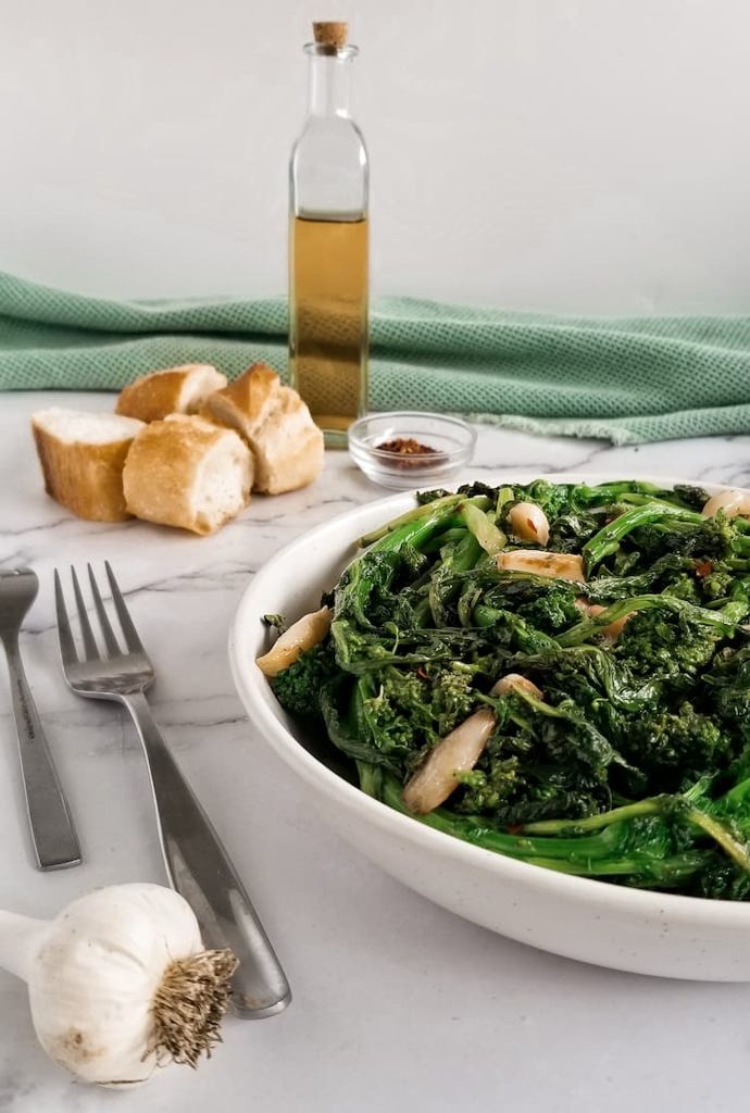 two forks next to a bowl of sauteed rapini with garlic, garlic bulb in the front, bottle of oil in the back next to 4 thick slices of bread and a small bowl of chili flakes