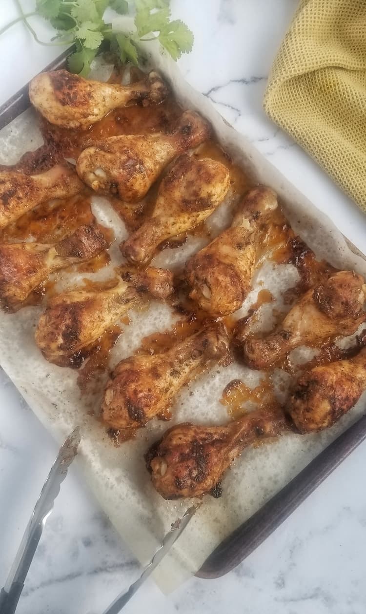 parchment lined sheet pan with 11 baked chicken drumsticks, fresh parsley at the back, yellow cloth on the side, tongs on the bottom left