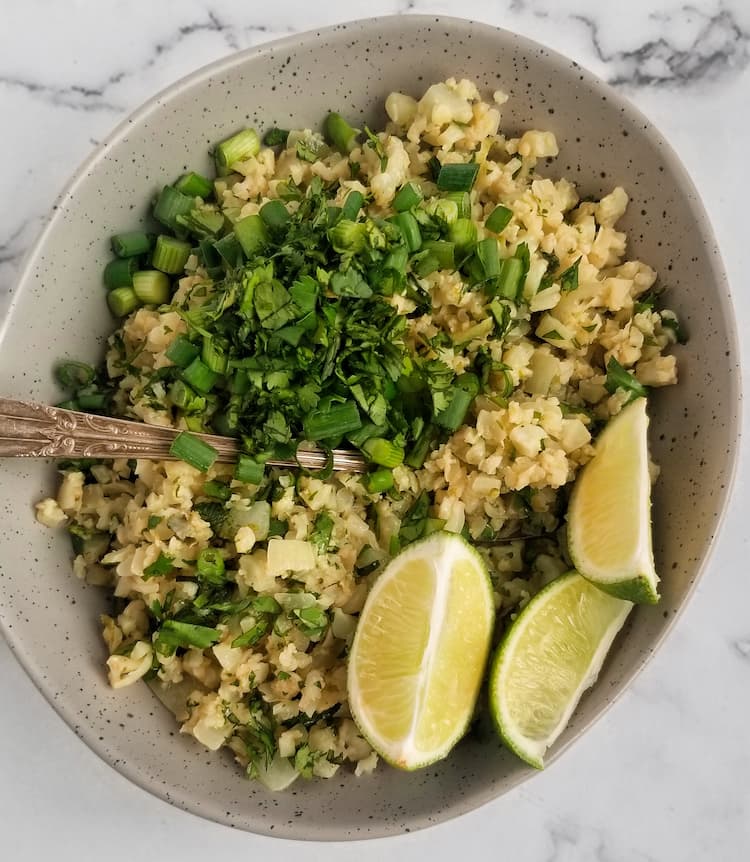 bowl of riced cauliflower, cilantro, lime wedges and chopped green onions, spoon in the bowl