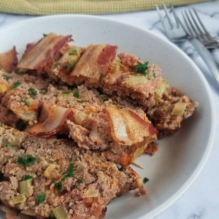 side view of bowl with 3 slices of keto meatloaf topped with cooked bacon and fresh chopped parsley, two silver forks on the right side, yellow cloth in the background