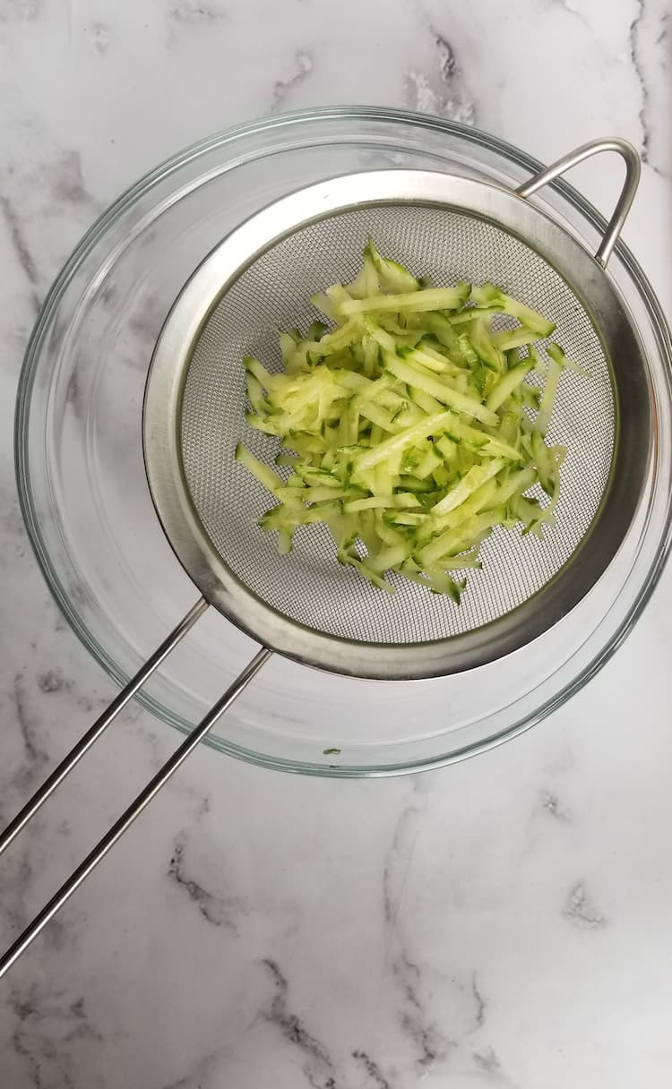 fine mesh strainer with shredded cucumber in it over a glass bowl