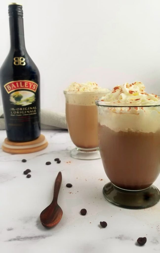 two clear cups filled with irish coffee with baileys, topped with whipped cream and cinnamon, bottle of baileys in the background, coffee beans and wooden spoon in the front