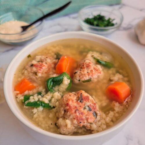 bowl of italian wedding soup with spinach, pasta, carrots, mini meatballs and broth. Bowls of parsley and cheese in the background