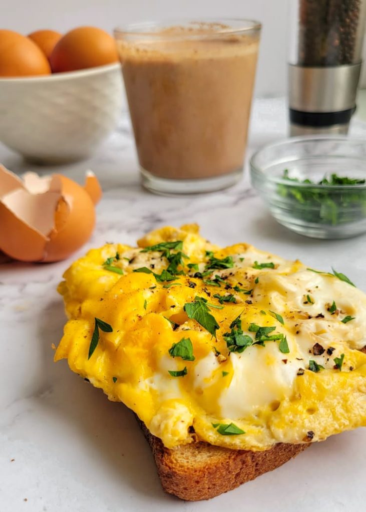 scrambled eggs on toast garnished with fresh parsley, fresh parsley in the background with a smoothie, a bowl of whole eggs, a peppermill and egg shells