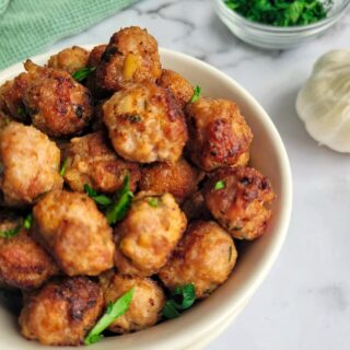 side view of bowl of polpette aka mini meatballs, fresh chopped parsley on top and in a bowl in the back, garlic bulb on the side