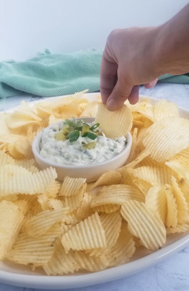 bowl of dill pickle dip topped with green onions and chopped pickles with some chips around it, hand dipping a chip into the dip