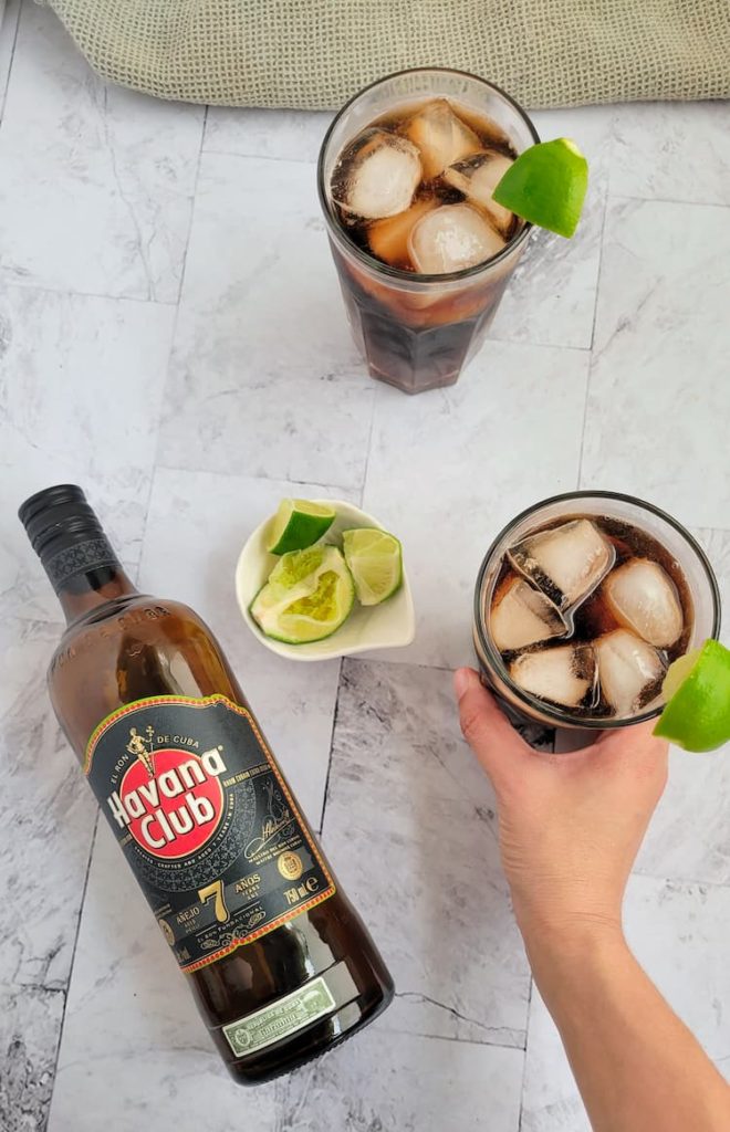 bottle of havana club 7 next to a small bowl of limes, hand holding a rum and coke with a lime wedge next to another glass of rum and coke