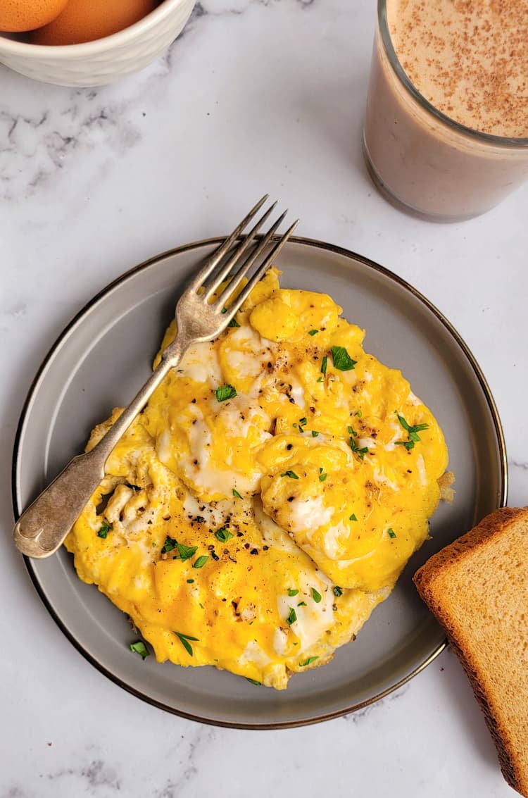 a plate of scrambled eggs garnished with chopped parsley with a fork and slice of toast, drink and whole eggs in the background