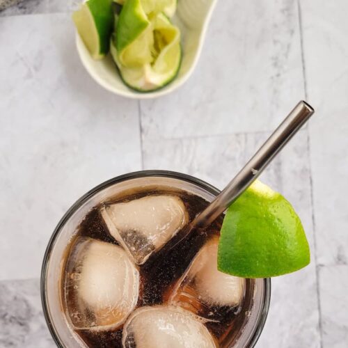 cuba libre with ice, a metal straw and a wedge of lime, other lime wedges in a bowl behind