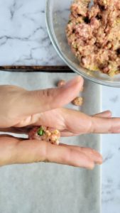 hand rolling meat into a meatball above a parchment lined baking sheet and a bowl of the meat mixture
