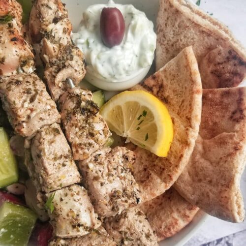 two pork souvlaki skewers in a bowl with some pita, lemon, greek salad and tzatziki sauce