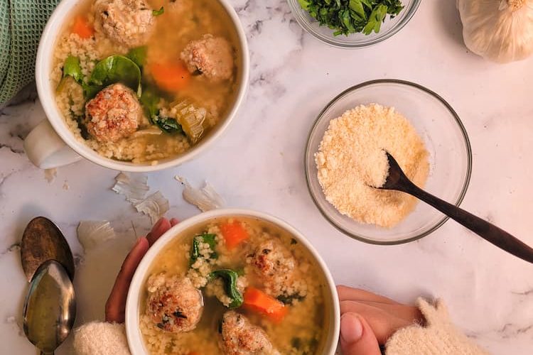 garlic bulb, chopped parsley and parmesan cheese in bowls surrounding two cups of italian wedding soup, two spoons on the side, hand holding one bowl