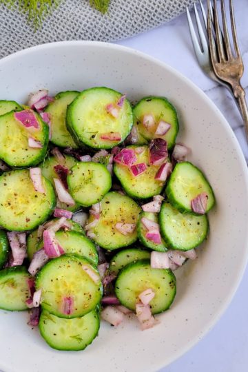 bowl of cucumber salad with dill with diced red onions, fresh dill in the background, two forks on the side