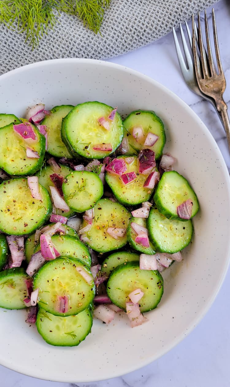 bowl of cucumber salad with dill with diced red onions, fresh dill in the background, two forks on the side