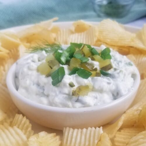 bowl of dill pickle dip topped with green onions and chopped pickles with some chips around it