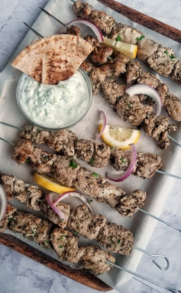 parchment lined baking sheet with pork souvlaki on metal skewers, tzatziki sauce with a triangle pita in it, lemon and red onion slices scattered about