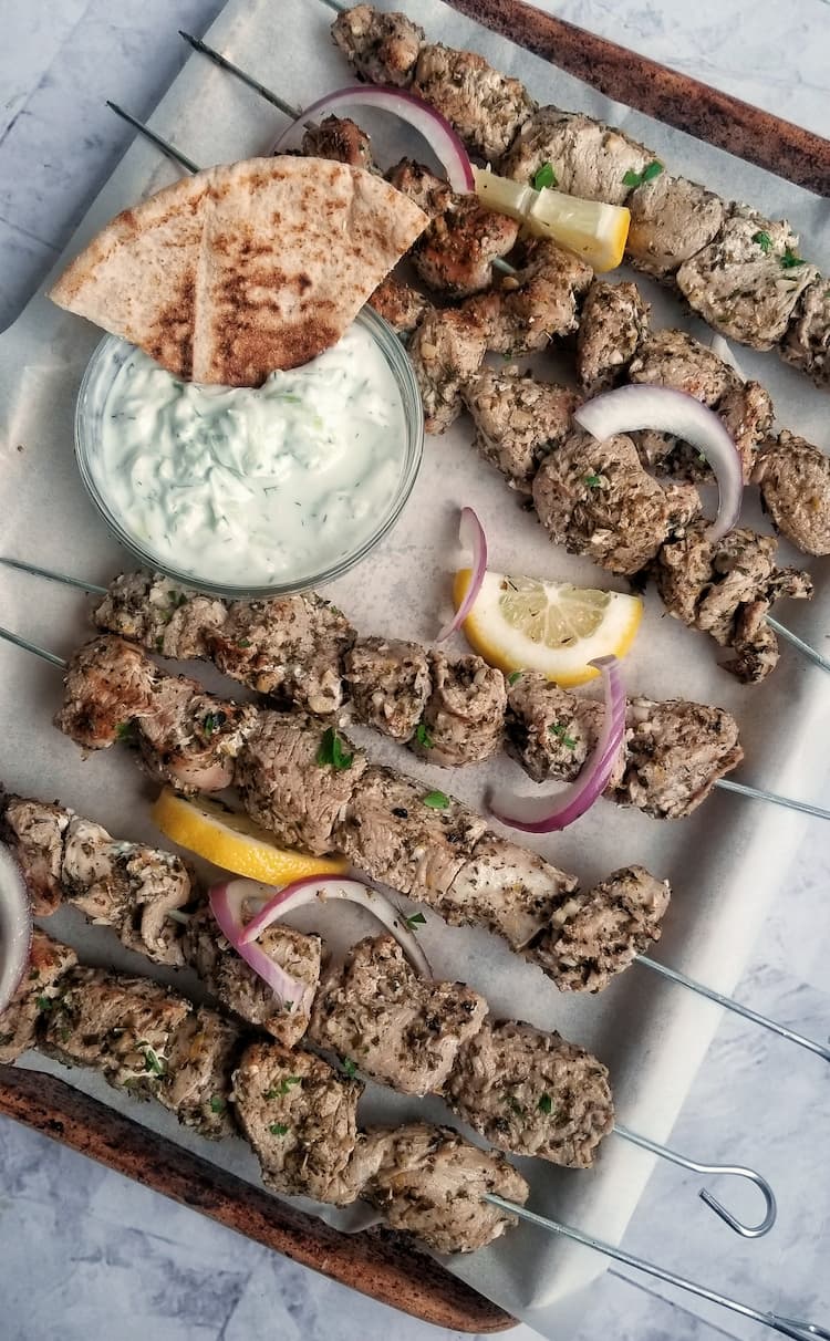 parchment lined baking sheet with pork souvlaki on metal skewers, tzatziki sauce with a triangle pita in it, lemon and red onion slices scattered about