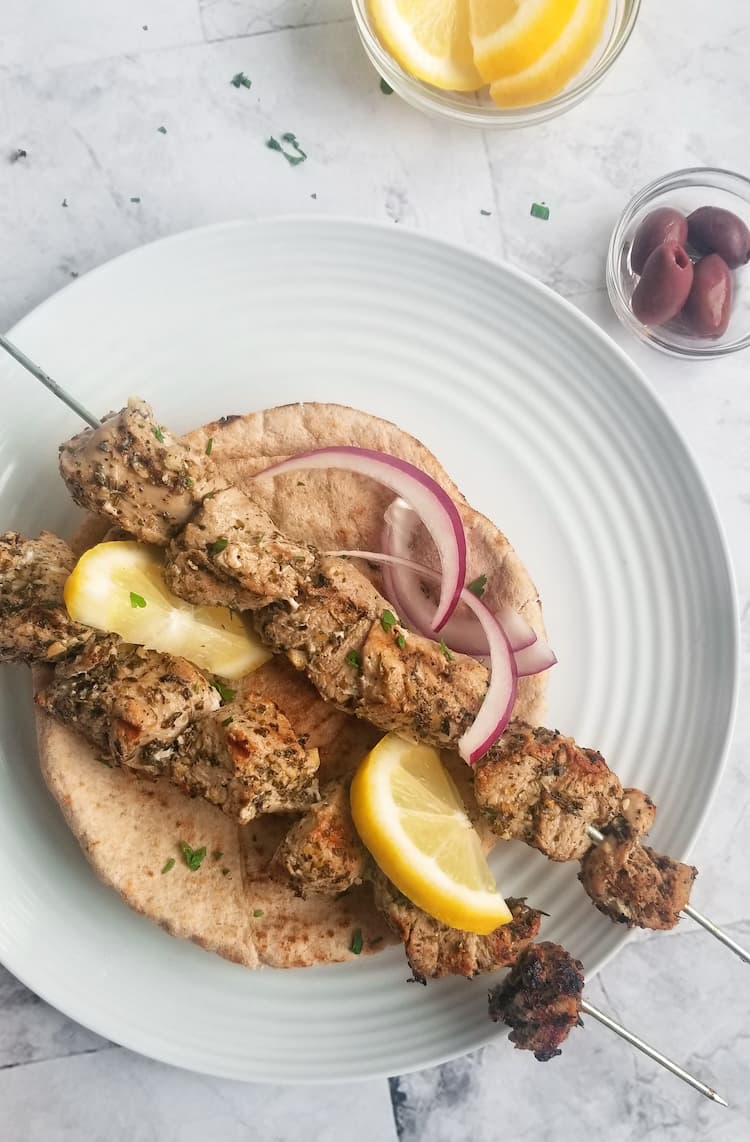 two skewered pork souvlaki on pita with sliced lemon and sliced red onion, small bowl of olives and another one of lemons on the side