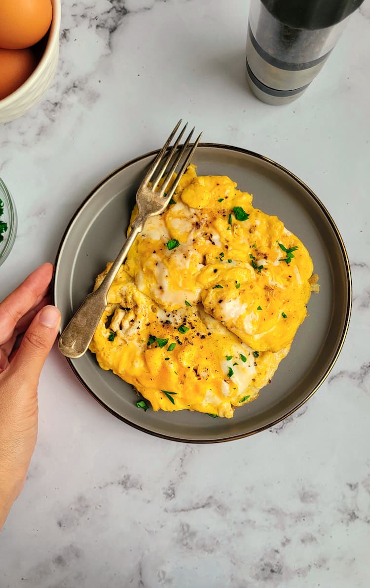 hand touching a plate with scrambled eggs garnished with parsley and a fork, whole eggs and peppermill in the background