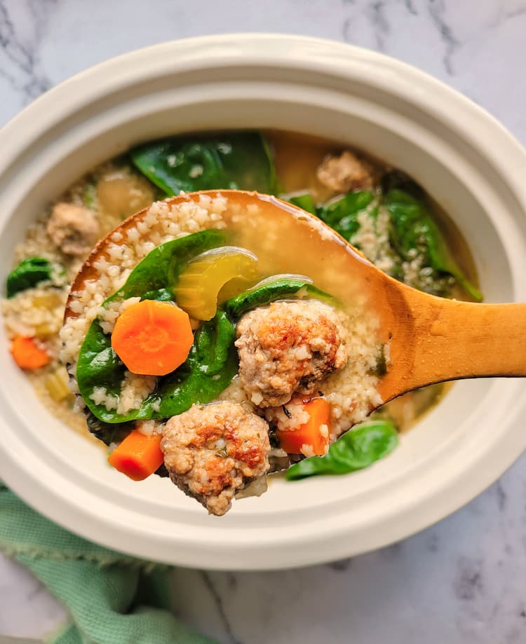 spoon holding up some italian wedding soup showcasing mini meatballs, spinach, celery, tiny pasta and carrots over a crockpot of the rest