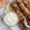 pork souvlaki on a plate next to a slice of lemon, a bowl of tzatziki and some triangular pita slices in the background