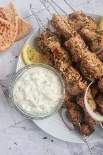 pork souvlaki on a plate next to a slice of lemon, a bowl of tzatziki and some triangular pita slices in the background