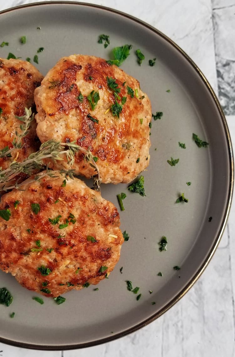 3 breakfast sausage patties on a plate with fresh chopped parsley and sprigs of fresh thyme