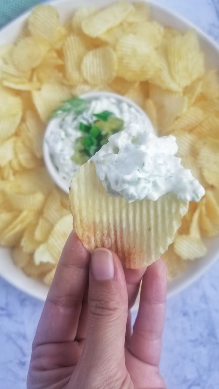 hand holding up a chip with white creamy dip on over a plate with more chips and dip