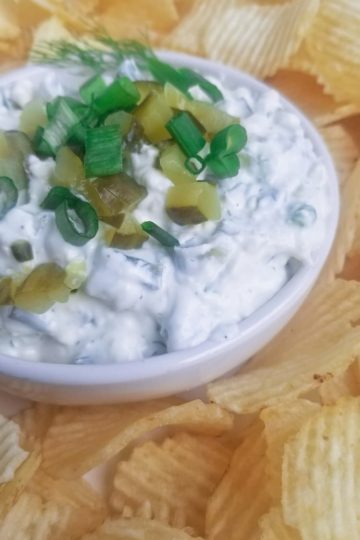 close up of bowl of dill pickle dip topped with green onions and chopped pickles with some chips around it
