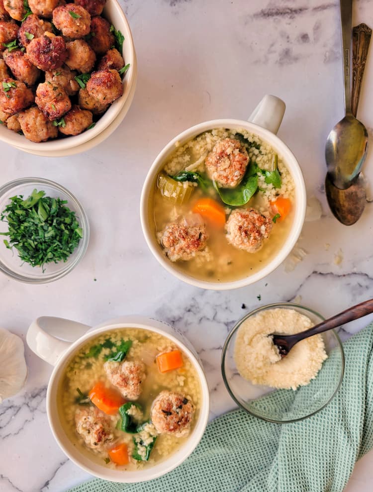 cooked meatballs, chopped parsley and parmesan cheese in bowls surrounding two cups of italian wedding soup, two spoons on the side