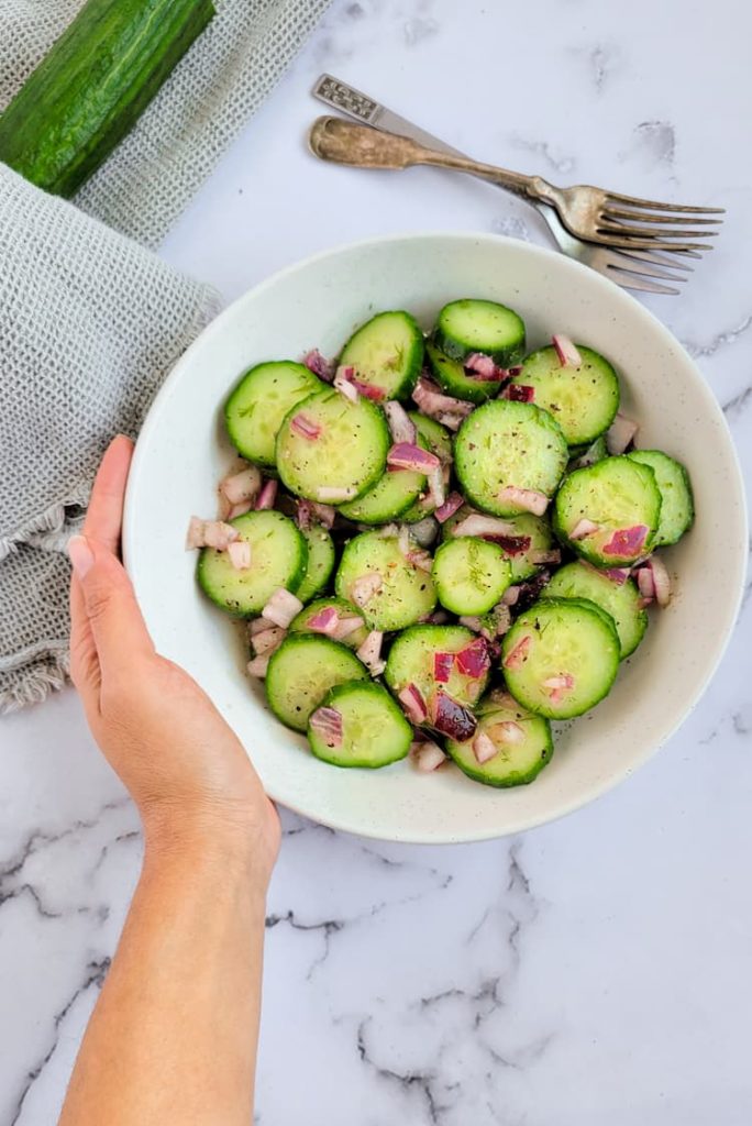 hand holding a bowl of cucumber salad with dill with diced red onions, cucumber in the background with 2 forks