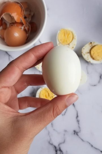 hand holding a whole hard boiled egg over some halves next to a bowl of shells