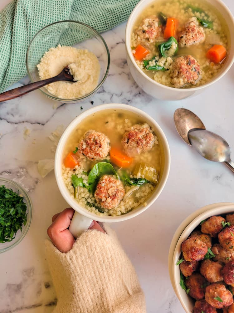 two bowls of italian wedding soup, hand on one bowl, bowl of meatballs at the bottom, bowl of fresh parsley and parmesan cheese on the other side, two spoons