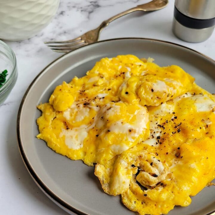 side view of scrambled eggs on a plate with salt and pepper, fork, whole eggs and peppermill in the background