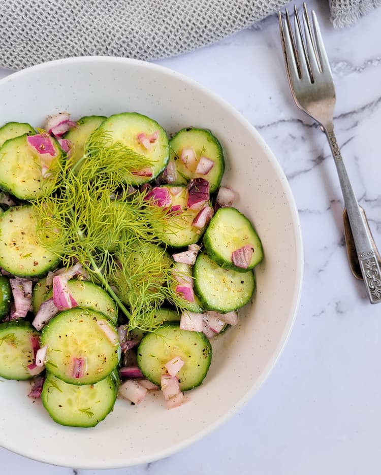 bowl of cucumber salad with dill with diced red onions, two forks on the side, fresh dill sprig on top of the salad