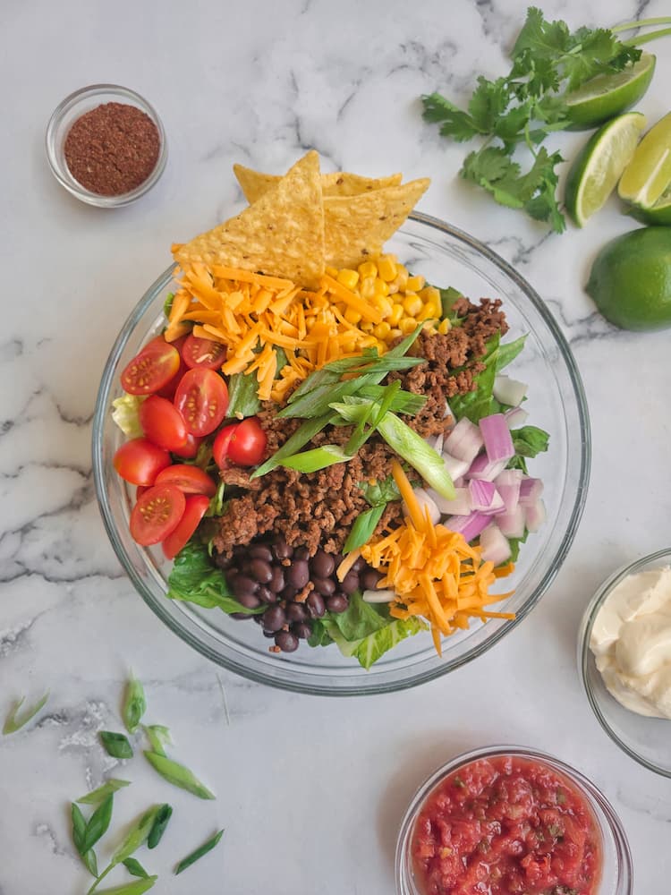 a taco salad in a large bowl with lettuce, corn, beans, onions, tomatoes, cheese, ground beef, tortilla chips. Sour cream, salsa, cilantro, lime wedges, sliced green onions and taco seasoning scattered around the salad