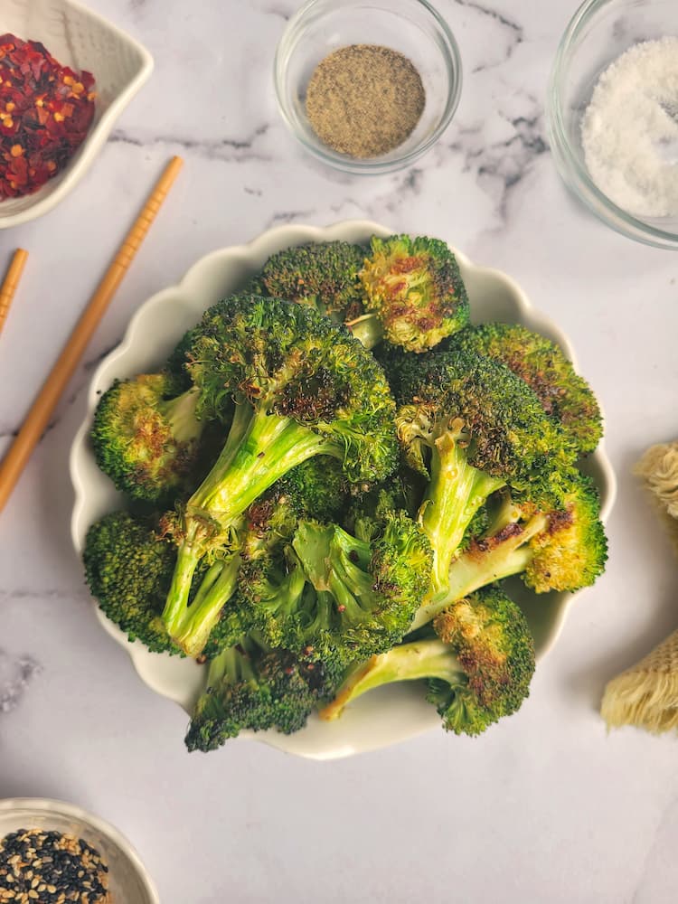 bowl of roasted broccoli, chopsticks on the side, ramekins of salt and pepper, chili flakes and sesame seeds surrounding