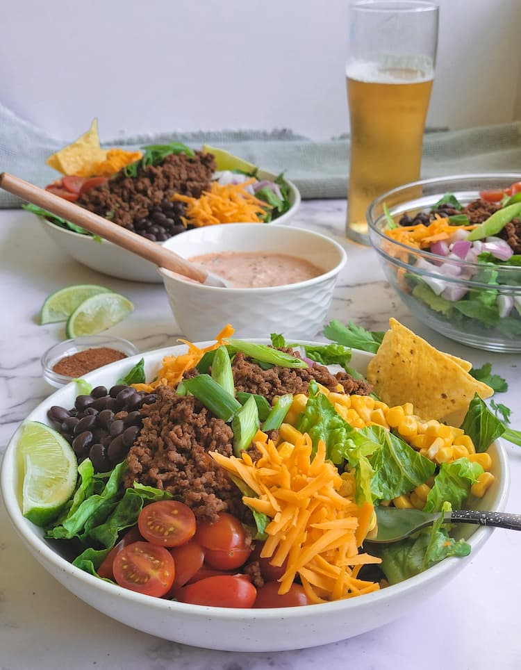 3 taco salads with tomatoes, cheese, lettuce, beans, ground beef, corn, onions, lime wedges. Taco seasoning, lime wedges, a beer and a bowl of salsa and sour cream also in the frame