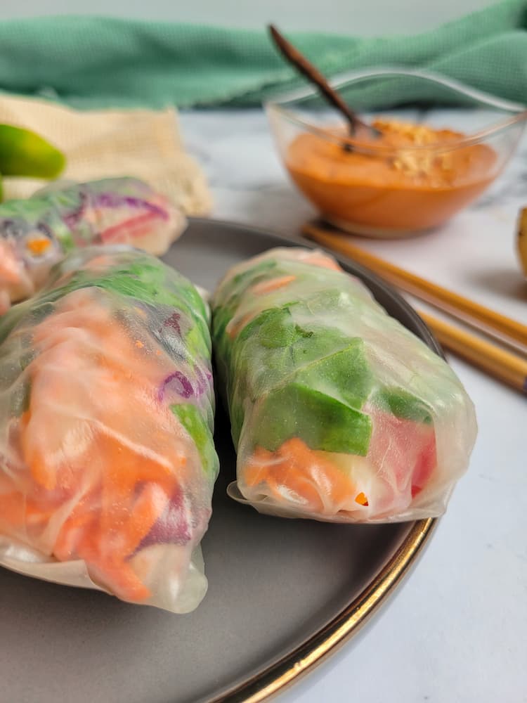 side view of fresh veggie spring rolls, chopsticks and a bowl of peanut sauce in the background