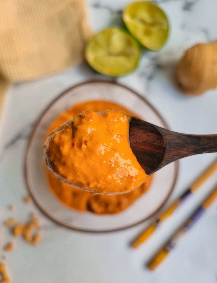 wooden spoon holding up some peanut sauce over a bowl of the rest, halved limes in the background with chopsticks and chopped peanuts