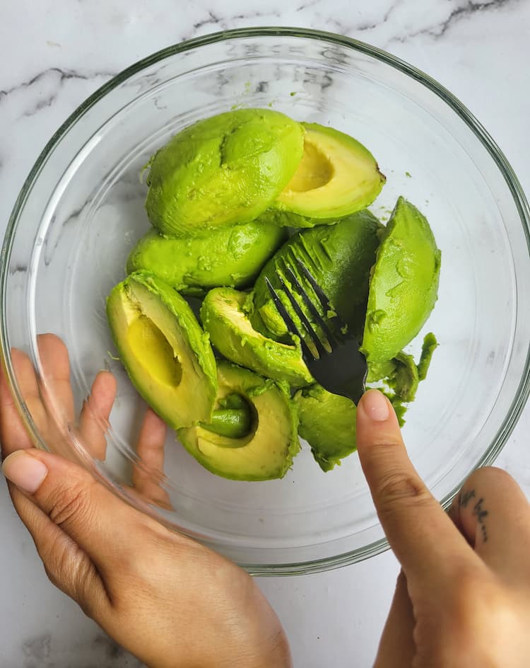 bowl of halved avocados, hand mashing one with a fork